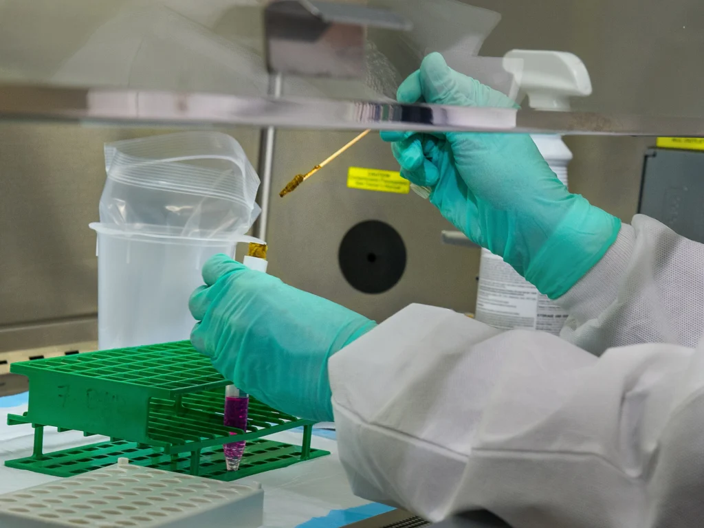 About us — A laboratory technician in a white coat and green gloves prepares samples using a pipette inside a sterile environment.